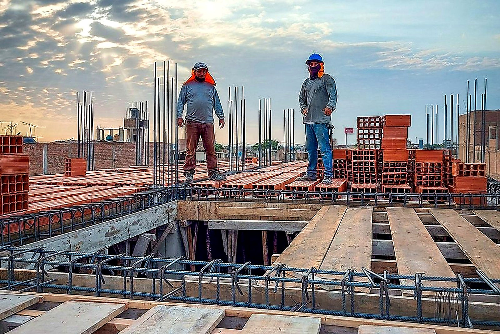 Free Two construction workers stand on a building site under a cloud-filled sky. Sunlight illuminates the scene. Stock Photo
